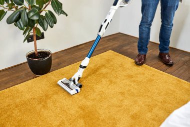 Kırpılmış adam, modern beyaz oturma odasında elektrikli süpürgeyle halı temizliyor.