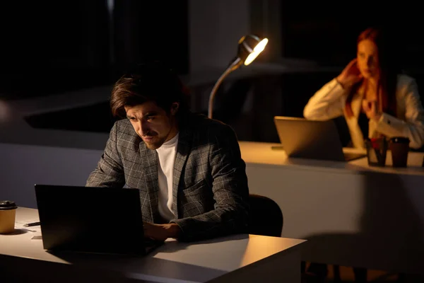 Kimerült, fókuszált fiatalember dolgozik távolról késő este az irodában számítógéppel — Stock Fotó