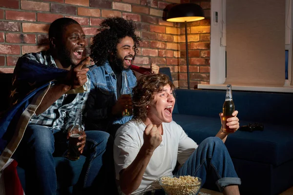 Amigos americanos bebiendo cerveza mientras animan a su equipo favorito en el campeonato de fútbol en la televisión — Foto de Stock