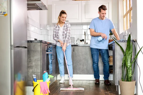 Limpieza de la casa, Pareja joven, En la cocina. Mujer limpiando piso con fregona, marido ayudando — Foto de Stock