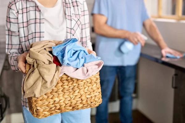 Mulher casualmente vestida com camisa quadriculada segurando cesta cheia de roupa suja precisando de lavagem, homem limpando cozinha no fundo — Fotografia de Stock