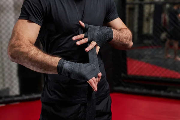 Обрізаний чоловік в чорному спортивному одязі готується до жорсткої боротьби, обгортаючи кулак в спортивних захисних бинтах — стокове фото