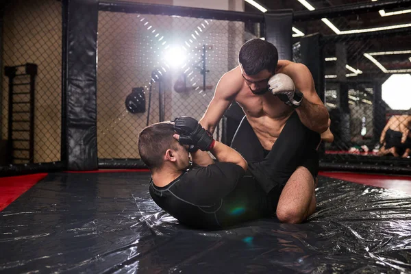 Shirtless muscular mma fighter in boxing gloves sitting above opponent while sportsman lying on floor resisting — стоковое фото