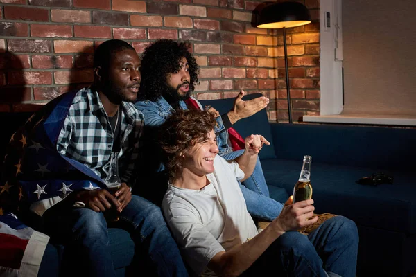 Jóvenes estudiantes viendo el deporte partido en casa sentado en el sofá. fanáticos animando a la selección nacional favorita, bebiendo cerveza — Foto de Stock