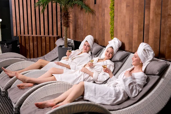 Young ladies enjoy drinking champagne at spa. Three beautiful women wearing bathrobes having cool resting, holidays — стоковое фото