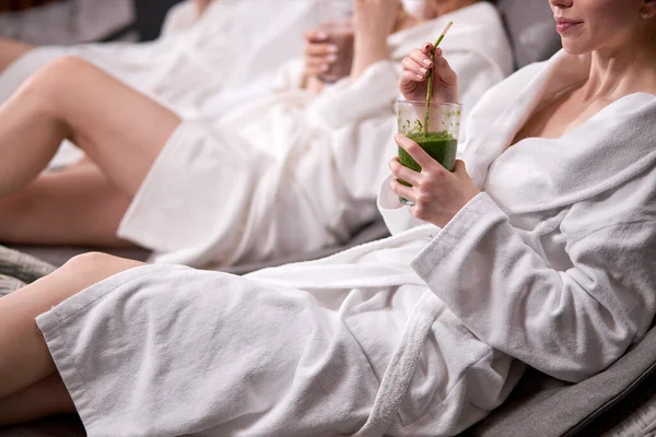 Close-up shot of young women wearing bathrobes drinking freshly squeezed juice while spending weekend in wellness center — Stock fotografie