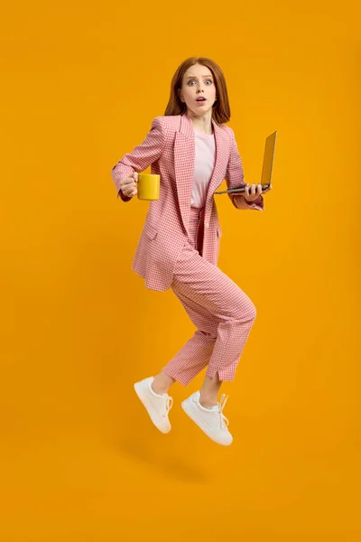 Red-haired woman jumping going using laptop web project isolated over yellow background