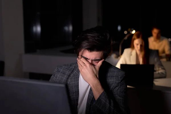 Νεαρός καυκάσιος μελαχρινός άνδρας με κοστούμι κάθεται τη νύχτα στο γραφείο, μάτια πληγωμένα από υπερκόπωση — Φωτογραφία Αρχείου