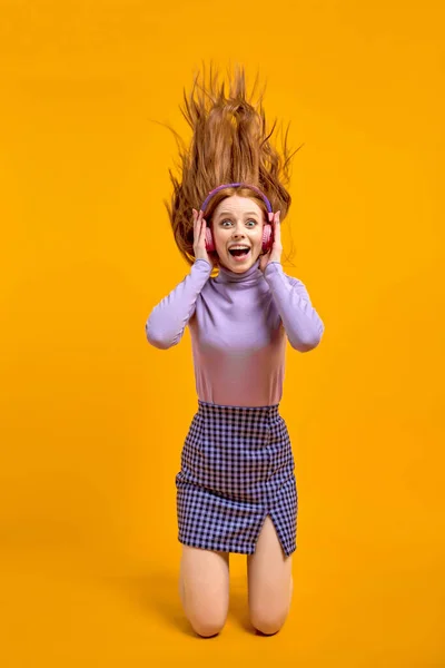 Эмоциональные молодые возбужденные женщины в наушниках с летящими волосами изолированы на желтый — стоковое фото