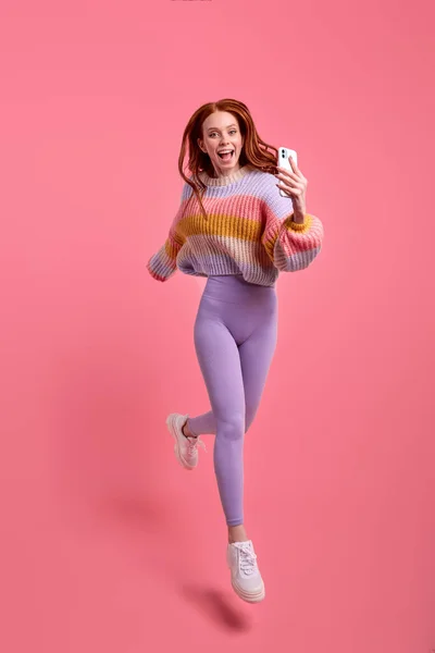 Caucasian Shocked redhead woman in casual clothes jumping and taking photo on smartphone — Stock Photo, Image
