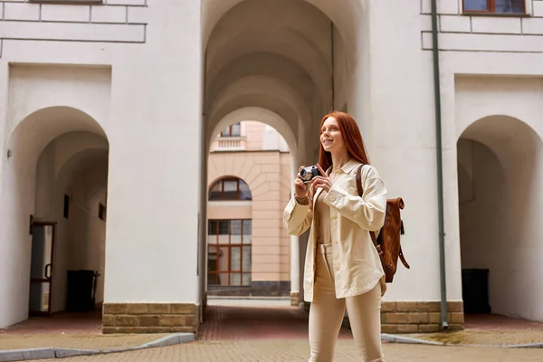 Vit rödhårig kvinna med retrokamera i händerna tittar på urban gammal arkitektur — Stockfoto