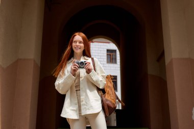 Avrupa 'nın eski şehrinde kamera kullanarak sokakta yürüyen kızıl saçlı kadın turist.