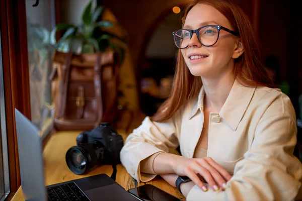 Reflexiva pelirroja fotógrafa femenina con la moderna tecnología portátil sentado en la cafetería — Foto de Stock