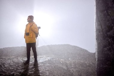 Dağın aşağısında, sisli havada yalnız yürüyen kameralı bir erkek.