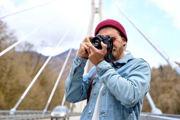 Hombre guapo en chaqueta de mezclilla caminando por el puente en el campo usando la cámara — Foto de Stock