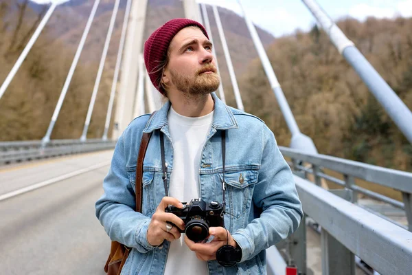 Hipster macho em jaqueta de ganga na ponte no campo usando câmera — Fotografia de Stock