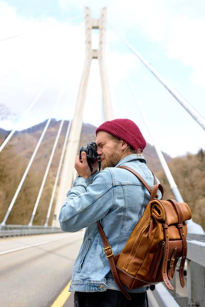 Przystojny hipsterski młody facet spacerujący po moście w obszarze miejskim robiący zdjęcia przed kamerą — Zdjęcie stockowe