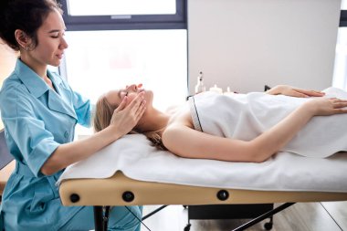 Yatakta yatan genç bir kadının yüzüne masaj yapan kadın masörün portresi.