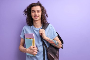 Yakışıklı, kıvırcık erkek, renkli renkli kitaplar. Kolejde ya da üniversitede okumaya başladığı için mutlu. Kameraya bakıyor.