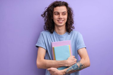 Kıvırcık beyaz adam, günlük tişörtlü kitaplarla stüdyoda mor duvar arkasında yalnız poz veriyor.