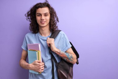 Kıvırcık saçlı, günlük kıyafetler giyen, sırt çantalı, mor arka planda saklanmış kitapları olan genç bir adam.