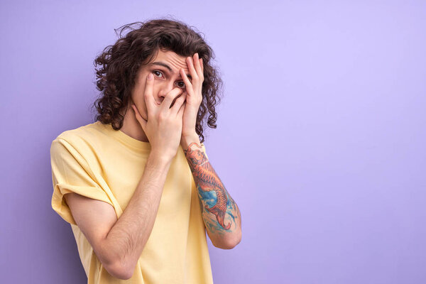 Young teen boy with curly hairstyle closing face with hands, looking through fingers with scared face expressions.