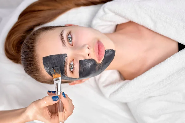 Žena relaxace s dřevěným uhlím obličejová maska, oříznuté profesionální kosmetička podání na obličej — Stock fotografie