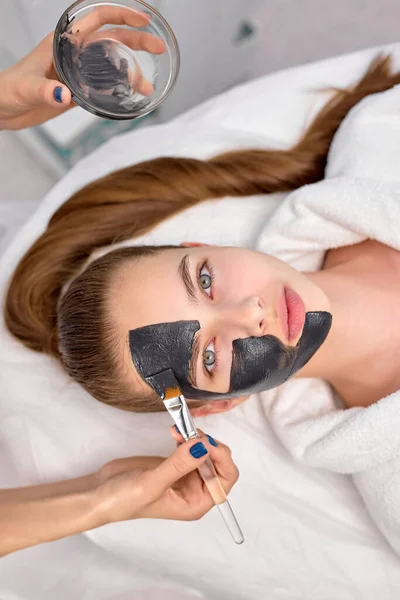 Procedura nakładania czarnej maski na twarz pięknej kobiety, widok z góry. Zabiegi spa — Zdjęcie stockowe