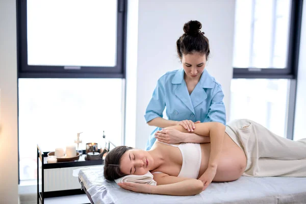 Professional massage therapist massages arm and shoulders of young pregnant woman