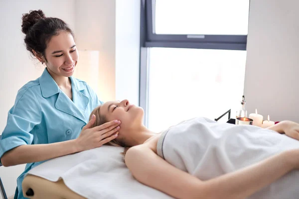 Zij uitzicht portret van vrouwelijke masseur doen massage op gezicht van jonge vrouw liggend op bed — Stockfoto