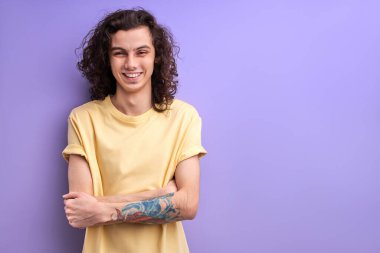 Uzun kıvırcık saçlı, gülen, kolları katlanmış, iyimser esmer adam.