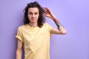 Uzun kıvırcık saçlı, beyaz, hoş bir genç. Kamerada gülümseyerek poz veriyor.