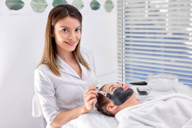 Genç kendinden emin kozmetik uzmanı müşterilerinin yüzüne fırça maskesi takıyor. Yan görünüm.