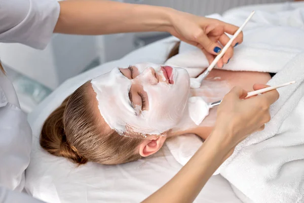 Młoda kobieta kosmetolog stosować białą maskę gliny na twarz kobiet.Widok z boku — Zdjęcie stockowe
