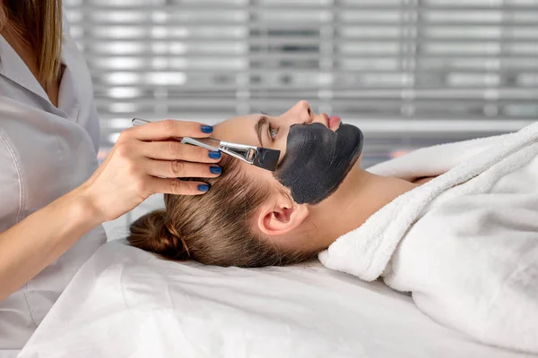 Professionelle Kosmetikerin, die eine Bürstenmaske auf das Gesicht ihrer Klienten aufträgt. Seitenansicht. — Stockfoto