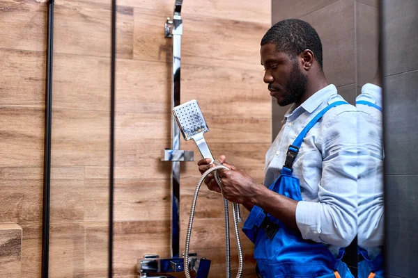 Афро-американский ремонтник в синем комбинезоне держит в руках ручку душа — стоковое фото