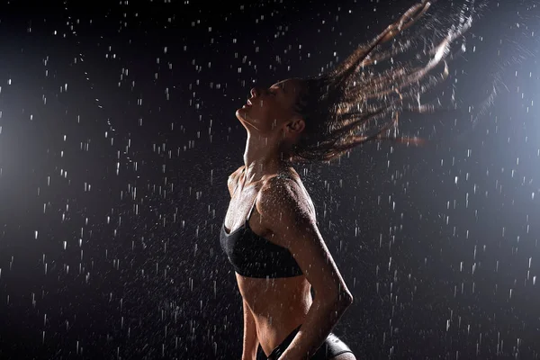 Mojada jovencita en traje de baño negro está bailando bajo la lluvia y salpicaduras, gotas de agua —  Fotos de Stock