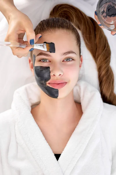 Hautpflege: Kosmetikerin cremt attraktive junge Frau mit schwarzer Gesichtsmaske ein — Stockfoto