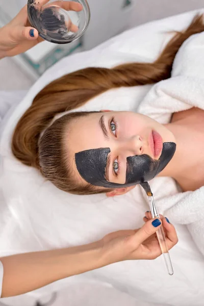 Top View On Cropped Kosmetolog použití černé masky na tváři krásné ženy — Stock fotografie