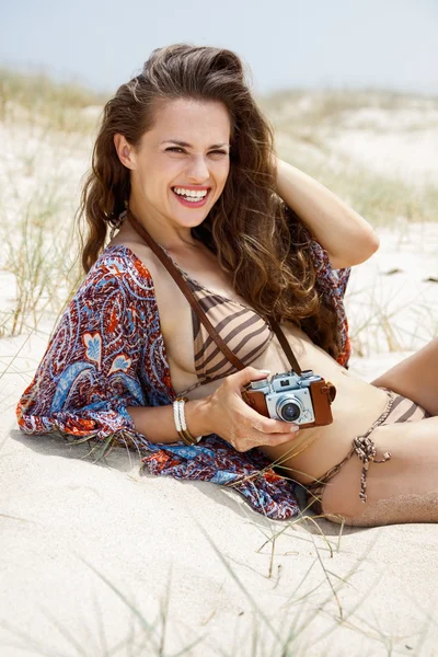 Smiling bohemian woman with retro photo camera sitting on beach — Stock Photo, Image