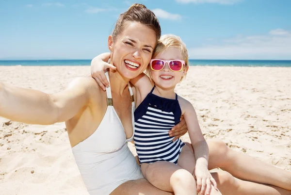 Sonrientes madre e hija en trajes de baño tomando selfies en la playa — Foto de Stock