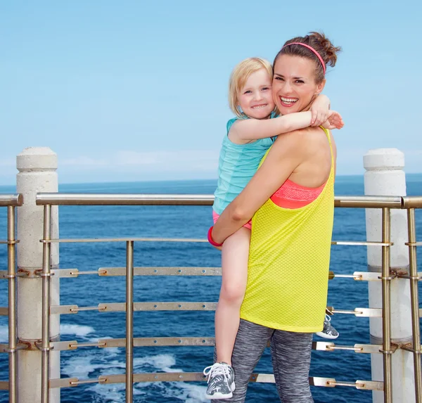 Smiling mother and child in fitness outfit hugging on embankment — Stock Photo, Image