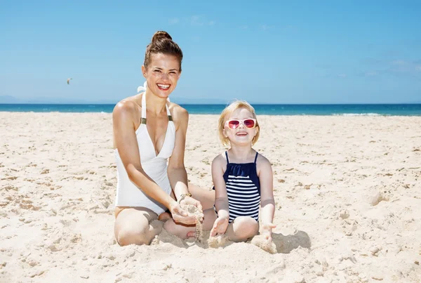 Lycklig mamma och flicka i badkläder på stranden leker med sand — Stockfoto