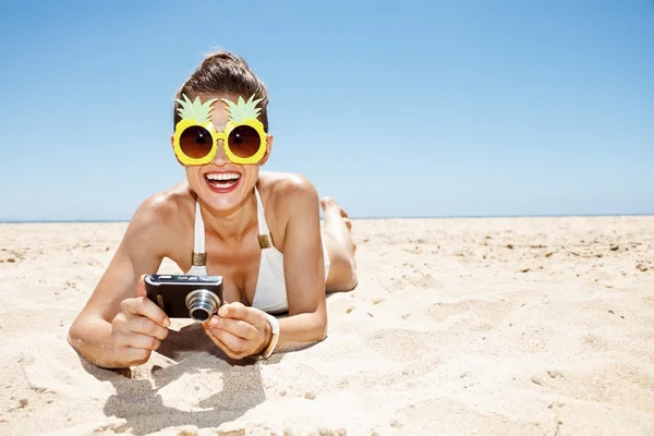 Smiling woman in pineapple glasses with photo camera at beach — Stock Photo, Image