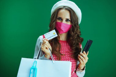 Pembe kolsuz gömlekli şık bir öğrencinin portresi. Pembe sağlık maskesi, alışveriş çantaları, dezenfektan ve kredi kartı satın alımı yeşil arka planda izole edilmiş bir akıllı telefondan yapılmış..