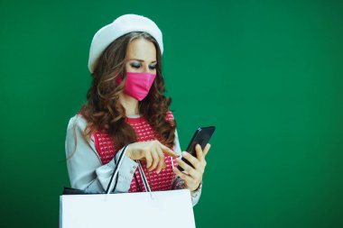 Koronavirüs salgını sırasında hayat. Pembe kolsuz gömlekli zarif bir öğrenci. Pembe tıbbi maskeli ve yeşil arka plandaki akıllı telefondan alışveriş torbaları alıyor..