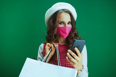 Beyaz bereli, pembe maskeli ve alışveriş çantalı gülümseyen modern kadın portresi yeşil bir telefondan e-ticaret sitesinden online alışveriş yapıyor..