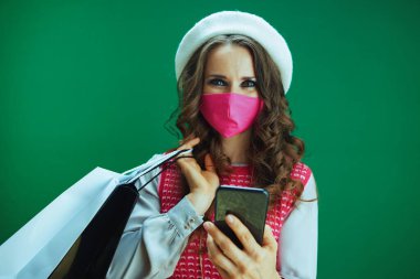 Koronavirüs salgını sırasında hayat. Pembe kolsuz gömlekli, pembe tıbbi maskeli ve yeşil arka planlı akıllı telefondan internetten alışveriş yapan mutlu, modern kadın alışverişçinin portresi..
