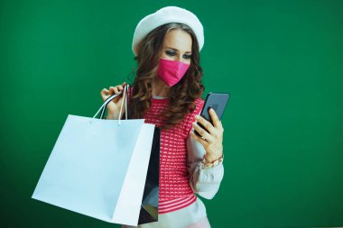 Covid-19 salgını sırasında hayat. Pembe tıbbi maskeli, alışveriş çantalı, beyaz bereli şık bir kadın yeşil arka plandaki akıllı telefondan online alışveriş yapıyor..