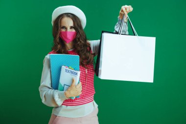 Koronavirüs salgını sırasında hayat. Pembe kolsuz gömlekli genç bir kadının portresi. Ders kitapları, defterler, pembe tıbbi maske ve yeşil arka planda alışveriş torbaları..